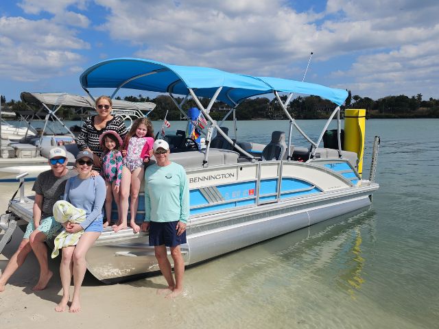 Pontoon boat anchored on a wide sandbar with two guests standing on shore, under a bright blue sky in Jupiter, Florida.